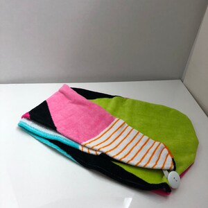 May include: A colorful hair towel with a button closure. The towel is made of a soft, absorbent material and features a geometric pattern in shades of pink, green, black, white, and orange.