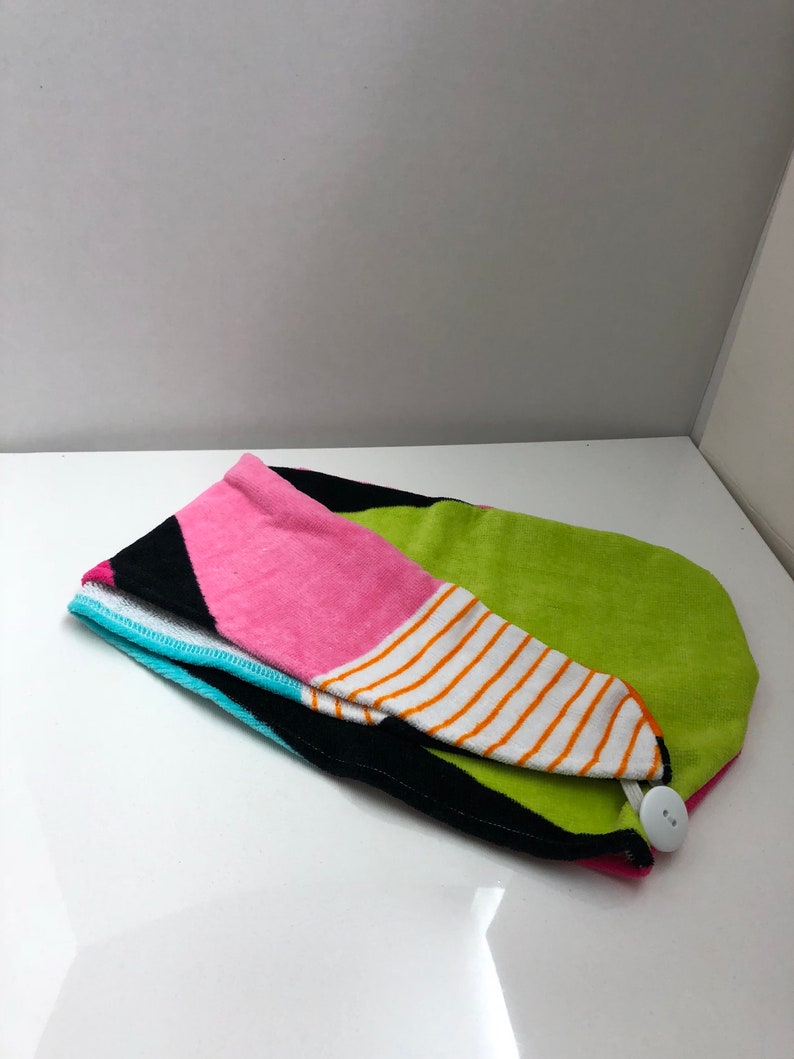 May include: A colorful hair towel with a button closure. The towel is made of a soft, absorbent material and features a geometric pattern in shades of pink, green, black, white, and orange.