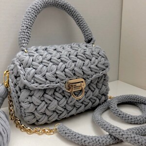 May include: A gray crocheted handbag with a gold chain strap and a matching tassel. The bag has a top handle and a flap closure with a gold clasp.