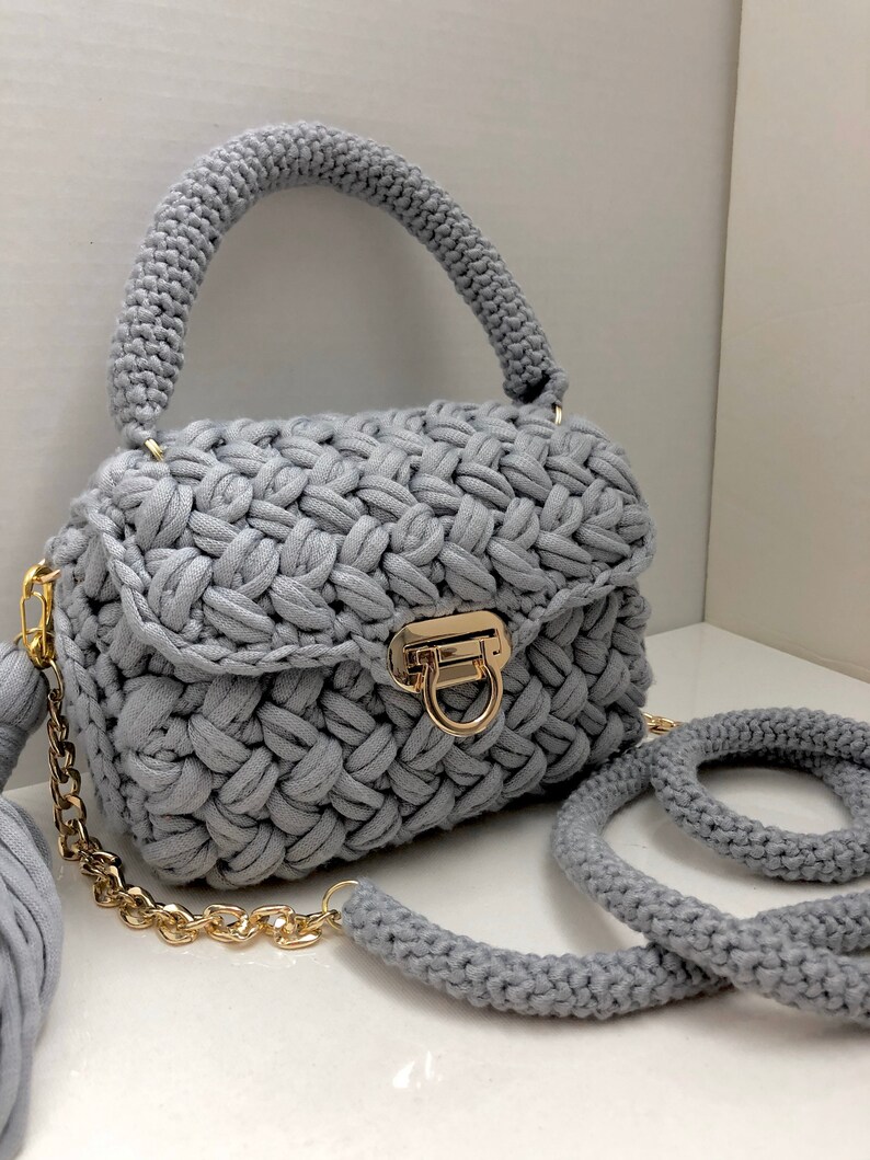 May include: A gray crocheted handbag with a gold chain strap and a matching tassel. The bag has a top handle and a flap closure with a gold clasp.