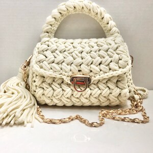 May include: A white crocheted handbag with a gold chain strap and a tassel. The bag has a top handle and a clasp closure.