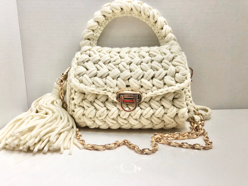 May include: A white crocheted handbag with a gold chain strap and a tassel. The bag has a top handle and a clasp closure.