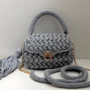May include: A gray crocheted handbag with a gold chain strap and a tassel. The bag has a flap closure with a gold clasp.
