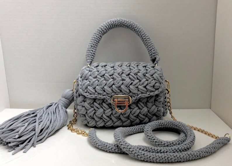 May include: A gray crocheted handbag with a gold chain strap and a tassel. The bag has a flap closure with a gold clasp.