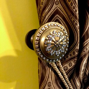 May include: A gold curtain tieback with a round, decorative finial covered in clear rhinestones. The tieback is attached to a brown and gold patterned curtain.
