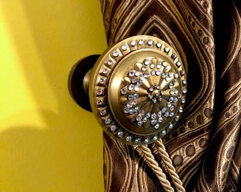May include: A gold curtain tieback with a round, decorative finial covered in clear rhinestones. The tieback is attached to a brown and gold patterned curtain.