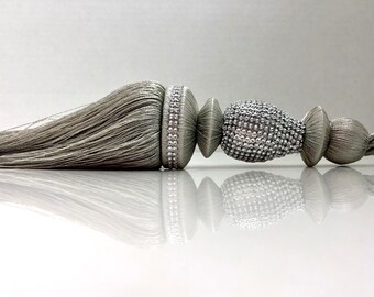 May include: A silver tassel tieback with a beaded design. The tassel is made of a soft, silky material and has a long, flowing fringe. The beaded design is intricate and adds a touch of elegance to the tieback.