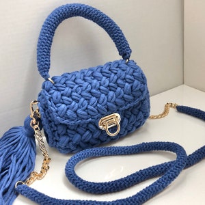 May include: A blue crocheted handbag with a gold chain strap and a tassel. The bag has a flap closure with a gold buckle.
