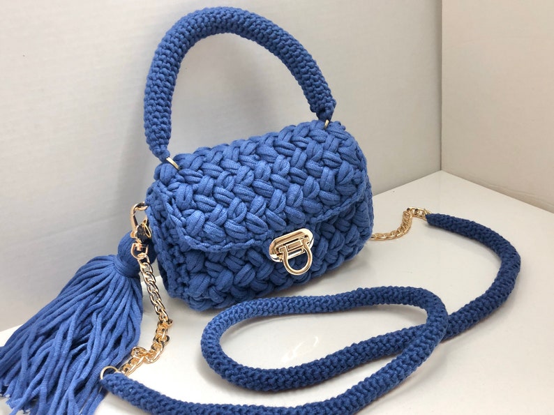 May include: A blue crocheted handbag with a gold chain strap and a tassel. The bag has a flap closure with a gold buckle.