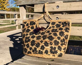 May include: A brown and black woven tote bag with a floral pattern. The bag is sitting on a wooden bench.