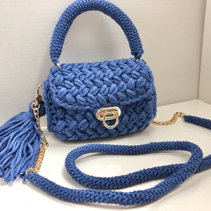 May include: A small blue crocheted handbag with a gold chain strap and a matching tassel. The bag has a top handle and a flap closure with a gold clasp.