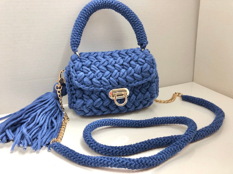 May include: A small blue crocheted handbag with a gold chain strap and a matching tassel. The bag has a top handle and a flap closure with a gold clasp.
