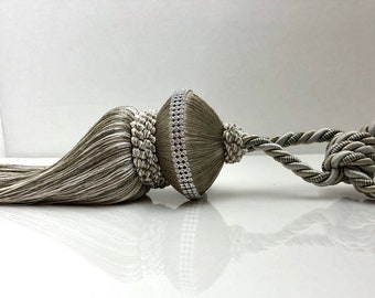 May include: A silver and gray tassel tieback with a decorative beaded trim. The tassel is made of a soft, woven fabric and has a long, flowing tail.