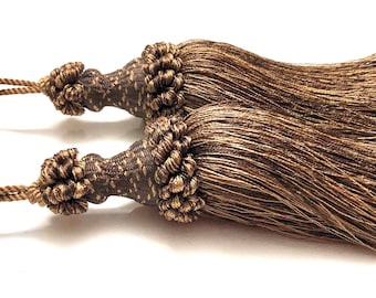 May include: Two decorative tassels with a brown and gold color scheme. The tassels have a knotted top and long, flowing strands. The cords are a matching brown color. These tassels are suitable for home decor and craft projects.