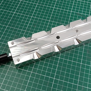 May include: A silver-colored metal injection mold featuring multiple elongated, tapered cavities for casting. The mold has a smooth, reflective surface and is attached to a solid black cylindrical handle extending from one end of the rectangular block.
