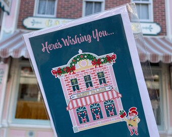 May include: A greeting card in a clear plastic sleeve featuring a dark teal background with the text Here's Wishing You... above an illustration of a pink brick building labeled Candy Palace, decorated with holiday garlands and a small elf figure.
