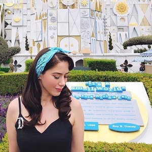 May include: A woman wearing a black dress and a blue bandana stands in front of the "it's a small world" ride at Disneyland. The ride is a colorful, whimsical structure with a sign that reads "it's a small world." The woman is smiling and looking at the camera.