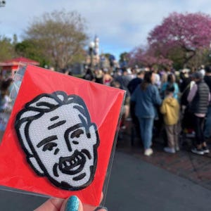 May include: A red packet holding a white and black embroidered patch featuring a cartoon face. The patch has a smiling expression and is surrounded by a black border. The background shows a crowd of people and trees.