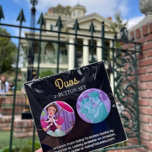 May include: A "Duos 2-BUTTON SET" featuring two round buttons with colorful illustrations. One button shows a woman with a parasol, the other a couple embracing. The background includes a historic building and a wrought iron fence.