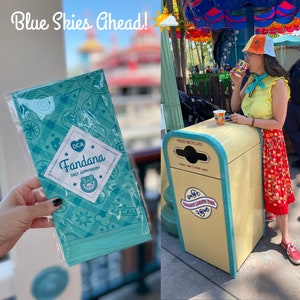 May include: A blue and white bandana with a cartoon bear and the text "DCA Fandana cala adventure" printed on it. The bandana is folded and packaged in a clear plastic bag.