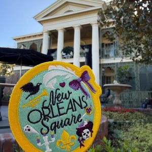 May include: A circular embroidered patch with a yellow border and a light blue center. It features the text New Orleans Square in black script, surrounded by a black bird, a white feather, a purple bow, a yellow trumpet, a ghost, a candelabra, a fleur-de-lis, and a skull with a purple hat.