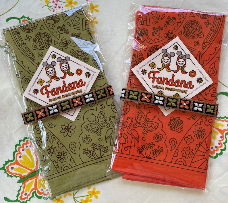 two colors of WDW golden anniversary shown in their packaging with true vintage 1970s flowered trim tied around them on true vintage 1970s fabric sheet with butterflies