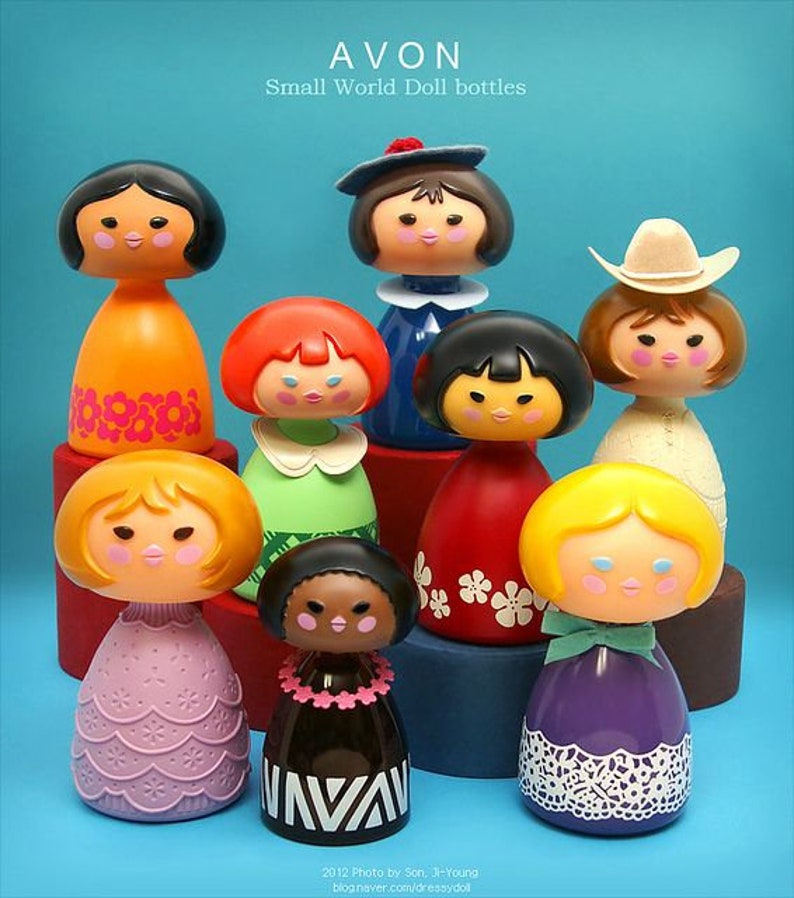 May include: A collection of eight colorful Avon Small World Doll bottles. The bottles are shaped like dolls with different hairstyles, clothing, and accessories. The bottles are arranged in a pyramid shape on a blue background.