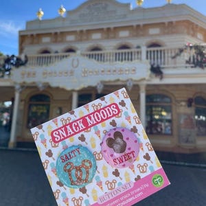 May include: A button set with the words "SNACK MOODS" on the top. One button is blue with a pretzel design and the word "SALTY". The other button is pink with a Mickey Mouse design and the word "SWEET".