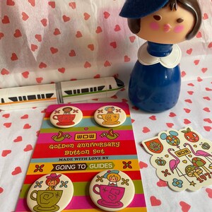 May include: A set of four colorful button pins featuring a cartoon character in a teacup. The buttons are packaged on a card with the text "WOW Golden Anniversary Button Set Made with Love by Going to Guides www.GoingToGuides.com".