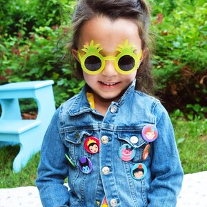 May include: A young girl wearing a blue denim jacket with various character buttons, yellow pineapple-shaped sunglasses, and a yellow polka dot dress.