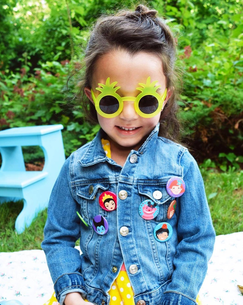 May include: A young girl wearing a blue denim jacket with various character buttons, yellow pineapple-shaped sunglasses, and a yellow polka dot dress.