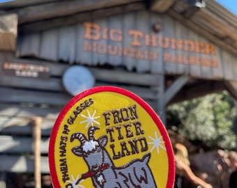 May include: A circular embroidered patch with a yellow background and a red border. It features a gray goat chewing on a red stick, with the text "FRONTIER LAND" and "HANG ON TO THEM HATS N' GLASSES" printed in black, accented by white star icons.