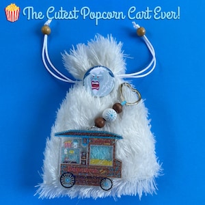 May include: A white fluffy bag with a popcorn cart charm, a round button with a cartoon yeti, and a snowflake charm. The text "The Cutest Popcorn Cart Ever!" is at the top. The bag has a drawstring closure with wooden beads.