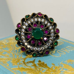 May include: A silver ring with a green stone surrounded by smaller white and pink stones. The ring has a detailed design with intricate swirls and filigree.