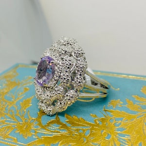 Könnte beinhalten: Ein silberner Ring mit einem großen, ovalen Amethyst-Edelstein, der in einem filigranen, komplizierten Design gefasst ist. Der Ring liegt auf einer blau-goldenen, gemusterten Oberfläche.