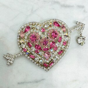 May include: A heart-shaped brooch with pink and clear rhinestones, featuring an arrow with a dangling heart charm.