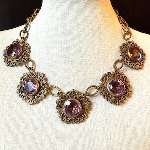 May include: A vintage necklace featuring five large, round, light purple gemstones set in ornate, filigree-style, gold-toned metal settings. The necklace has a chain with connecting links. The necklace is displayed on a white surface.