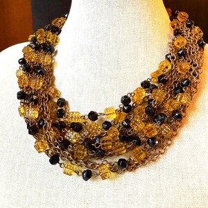 May include: A multi-strand necklace featuring alternating strands of amber-colored, square-cut beads and black, round beads. The beads are strung on gold-toned chains, creating a layered effect. The necklace is displayed on a white mannequin.