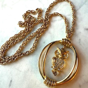 May include: Gold-toned necklace featuring a pendant with an owl design. The owl is embellished with clear rhinestones and hangs within a double-hoop frame. The chain is a textured, rope-style design.