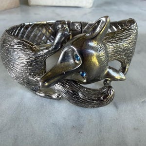 May include: A silver-toned metal hinged cuff bracelet featuring a sculpted fox design. The fox has textured fur detailing and two small, bright blue inset stones for eyes. The band of the bracelet displays a cross-hatched, woven-style pattern.