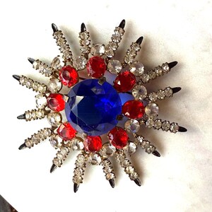 May include: A starburst brooch with a large blue central stone, red and clear gemstones, and silver-toned metal accents. The starburst points have black tips. A vintage-style brooch, suitable for adding a touch of glamour.