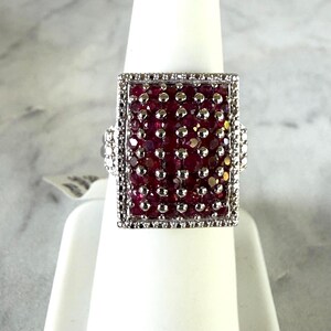May include: A rectangular silver ring featuring a cluster of deep red, faceted stones set in a grid pattern. The central stones are interspersed with small silver beads, and the entire rectangular face is bordered by a frame of tiny, clear stones.