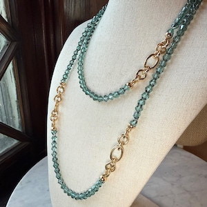 May include: A long, layered necklace featuring light blue faceted beads and gold-tone chain links. The necklace is displayed on a white mannequin bust, showcasing its length and design. The beads have a subtle sparkle, adding to the necklace's elegance.