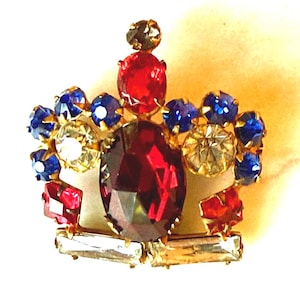 Op de afbeelding: Een vintage kroonvormige broche met een goudkleurige zetting. De broche heeft een grote, dieprode centrale steen, omringd door kleinere rode, blauwe en heldere edelstenen. Het ontwerp is sierlijk en gedetailleerd, met een klassieke, koninklijke uitstraling.