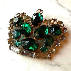 May include: Vintage brooch with a gold-tone setting. The brooch features a heart-shaped design with large, faceted emerald green and clear crystal stones. The stones are surrounded by smaller, clear crystals.