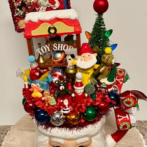 May include: A Santa Claus-shaped mug overflowing with Christmas decorations. It features a miniature toy shop, a small Christmas tree, ornaments, and a sign that reads "When Santa Gets Your Letter." The mug is on a wooden surface.