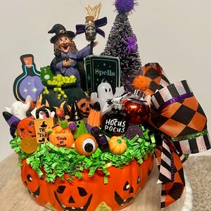 Puede incluir: Centro de mesa temático de Halloween con una base de calabaza naranja tallada, llena de decoraciones festivas. Incluye una figura de bruja, un árbol negro, fantasmas, calabazas y un adorno de "Hocus Pocus". El arreglo está acentuado con papel verde y un lazo a cuadros.