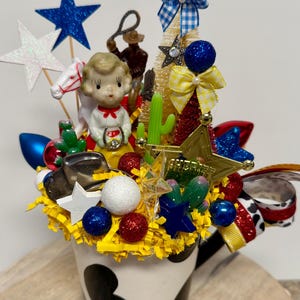 May include: A white mug with black cow spots filled with yellow paper shreds, a vintage-style figurine, a miniature cowboy, a plastic horse, green cacti, a gold sheriff star, glittery stars, red and blue spheres, and patterned ribbons.