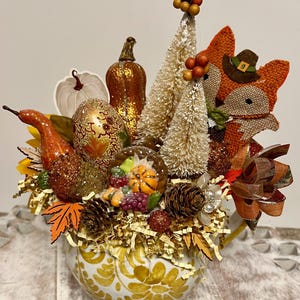 Może przedstawiać: Dekoracyjna aranżacja w jesiennym stylu w ceramicznym pojemniku z żółtym i białym wzorem. Aranżacja zawiera dynie, szyszki, jagody i małe drzewka szczotkowe. Kolorystyka jest ciepła, z odcieniami pomarańczu, brązu i złota.