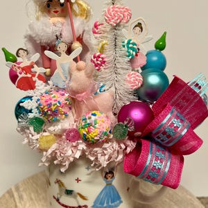 Może przedstawiać: Kapryśna aranżacja w dekoracyjnym kubku, z lalką wróżki, miniaturową choinką, ozdobami i słodyczami. Kubek zdobią ilustracje dziewczynki i konika na biegunach. Różowa kokarda z niebieskimi akcentami dodaje świątecznego charakteru.